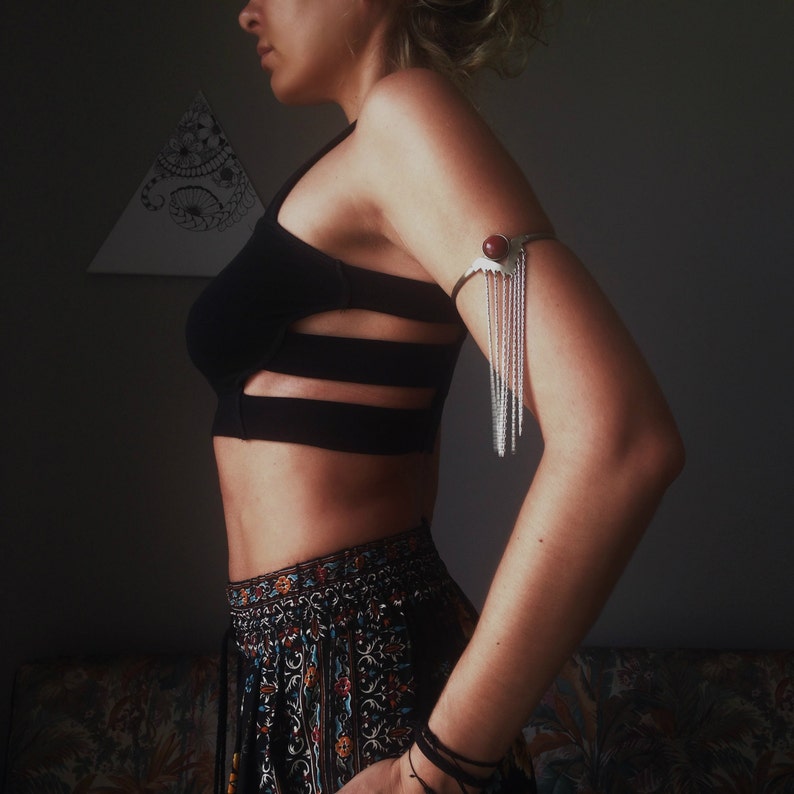 May include: A woman wearing a black crop top with cutouts and a silver arm cuff with a red stone. She is also wearing a patterned skirt and bracelets.