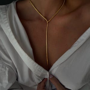 May include: A gold-toned snake chain necklace featuring a lariat design with a long, slender drop that extends downwards from a central knot. The chain has a smooth, flexible texture and a polished finish, resting against a white button-down shirt.