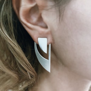 May include: A pair of silver geometric earrings with a triangular top and a curved bottom. The earrings are worn by a person with blonde hair.