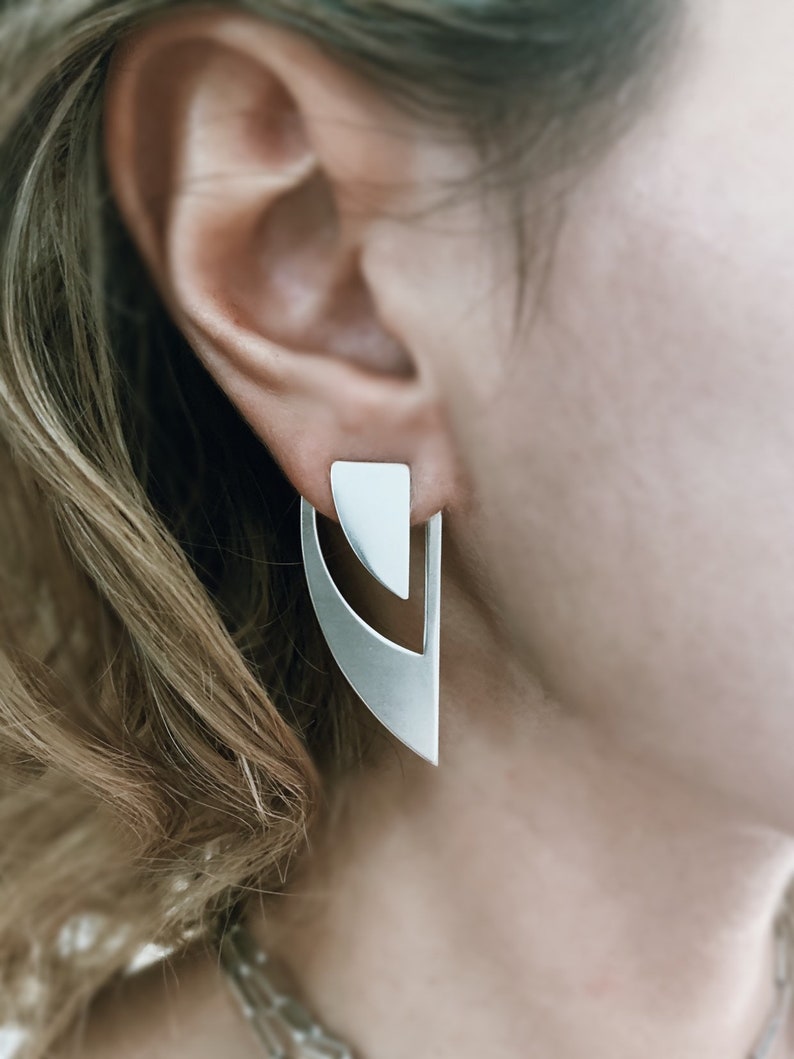 May include: A pair of silver geometric earrings with a triangular top and a curved bottom. The earrings are worn by a person with blonde hair.