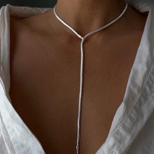 Könnte beinhalten: Eine silberfarbene Schlangenkette als Lariat-Halskette mit einer flexiblen, glatt strukturierten Kordel, die um den Hals verläuft und mittig auf der Brust herabhängt, wobei sie in einer schlanken, spitz zulaufenden Metallspitze endet.