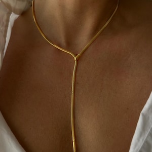 Könnte beinhalten: Eine goldfarbene Halskette mit Y-förmigem Design. Die Halskette hat eine zarte Kette, die einen langen, schlanken Anhänger bildet. Die Halskette wird über einem weißen Hemd getragen.
