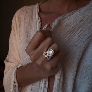 Op de afbeelding: Een zilveren ring met een waaiervormige bovenkant met stralende lijnen en twee symmetrische spiraalvormige krullen aan de basis. De ring wordt aan een vinger gedragen tegen een wit, gestructureerd kledingstuk met franjes aan de rand.