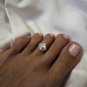 Puede incluir: Un anillo para el dedo del pie de tono plateado que presenta una pieza central prominente en forma de pirámide cuadrada. El anillo se lleva en el segundo dedo del pie y muestra un acabado metálico liso con líneas geométricas.