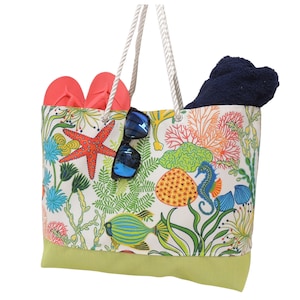May include: A large beach tote bag with a marine life design. The bag features a white background with colorful coral, fish, and seahorses. It contains a navy blue towel, a pair of coral flip-flops, and blue sunglasses. The bag has a green bottom and rope handles.
