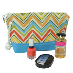 May include: A colorful chevron patterned zippered pouch with a blue bottom, a red nail polish bottle, a clear glass bottle of perfume with a pink lid, and a black eyeshadow palette.