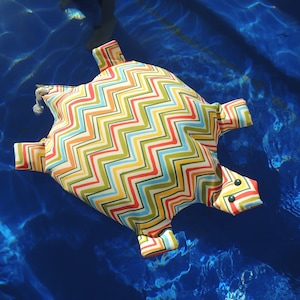May include: A colorful zig-zag patterned turtle-shaped pool float with black eyes. The float is floating in a blue pool.