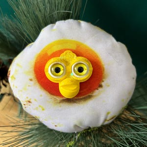 May include: A whimsical, handmade egg-shaped ornament with a yellow, bird-like face. The egg white is white with brown spots, and the yolk is yellow and orange. The ornament is decorated with black eyes and is set against a backdrop of green pine needles.