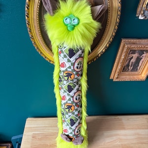 May include: A neon green plush creature with brown ears and feet, and a patterned body. The body features a repeating pattern of a silhouette, footprints, and the text "????". The creature is mounted on a gold framed oval.
