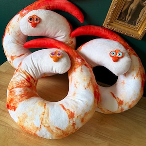 May include: Three plush shrimp pillows with white bodies, orange and red markings, and red tail fins. Each pillow has a whimsical face with orange beaks and googly eyes. The pillows are arranged on a wooden surface.