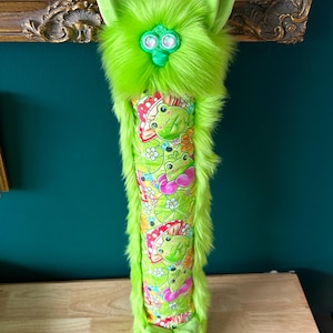 May include: A whimsical, lime green plush creature with large ears and a furry body. The central section features a colorful pattern of frogs, flowers, and bows. The toy is designed to stand upright.