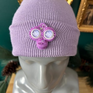 May include: A lilac-colored beanie hat with a decorative purple embellishment. The embellishment has two circular eye-like features with a glittery finish and a small, stylized face. The hat is displayed on a mannequin head.