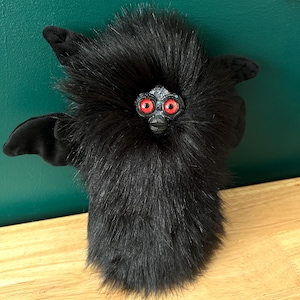 May include: A plush, black creature with red eyes and a beak-like nose. The creature has a fluffy, furry body, small wings, and ear-like appendages. It is a whimsical, handmade toy.