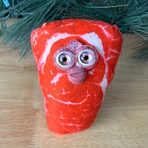 May include: A plush toy resembling a cut of raw meat, with a face featuring large, glittery eyes and a small nose. The toy is red with white marbling, and is positioned in front of a green, artificial Christmas tree branch.