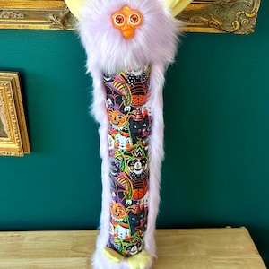 May include: A whimsical, plush Halloween-themed Furby-inspired creation. It features a lavender fur exterior, yellow ears and feet, and a patterned cylindrical body with Halloween characters. The design includes orange, black, and white elements, creating a playful, festive aesthetic.
