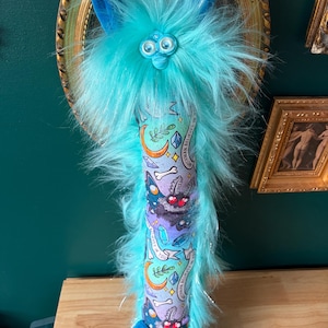 May include: A whimsical, plush creature with turquoise fur and a patterned body. The creature has large, round eyes and a beak-like nose. The body is wrapped in fabric with a repeating pattern of moons, crystals, and text. The creature's ears and feet are blue.