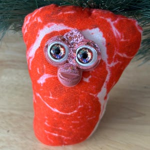May include: A novelty Christmas ornament shaped like a piece of raw steak. The red fabric has white marbling, and the ornament features large, glittery eyes and a pink face. Green pine needles are visible at the top.