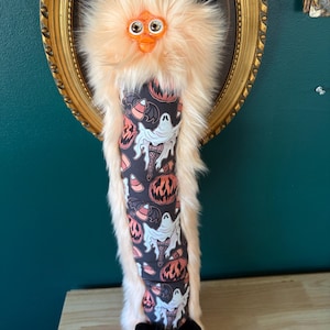 May include: A plush, Halloween-themed creature with peach-colored fur, black ears, and an orange beak. The body features a black fabric with white ghosts, orange pumpkins, and candy corn. The creature is mounted in a gold-framed oval.