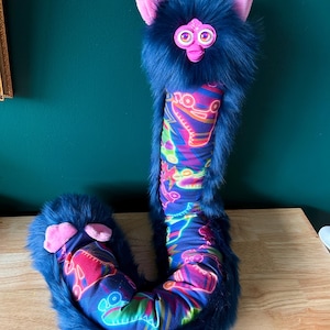 May include: A plush, whimsical creature with a dark blue, furry exterior. It features pink ears, a pink face, and a patterned body with neon-colored designs. The toy is designed to resemble a Furby.