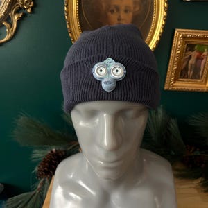 May include: Navy blue knit beanie with a decorative, light blue and white brooch. The brooch has a unique, abstract design with two circular elements. The beanie is displayed on a mannequin head.