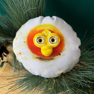May include: A plush, egg-shaped ornament with a yellow and orange yolk design. The ornament features a yellow beak and glittery eyes, resembling a cartoon character. It is surrounded by green pine needles.