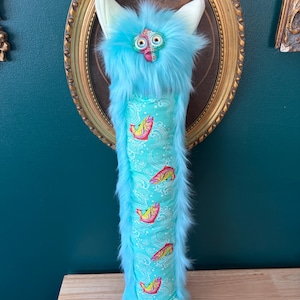 May include: A whimsical, plush creature with a turquoise body and fluffy fur. It has a patterned belly with colorful fish and wave designs. The creature has large, expressive eyes and a unique, playful appearance.