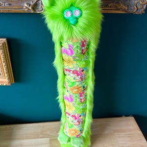 May include: A neon green plush creature with large eyes and ears. The body is a vibrant floral pattern with mushrooms and flowers. The creature has a furry texture and is standing on a wooden surface.