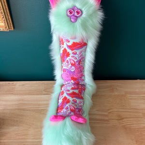 May include: A whimsical, plush creature with a mint green fur body, pink ears, and a patterned fabric midsection. The fabric features a floral design with red and pink accents. The creature has a purple face and pink feet, creating a playful and unique design.