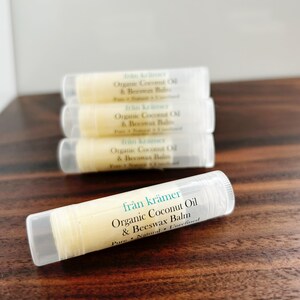 Pu&ograve; includere: Quattro tubetti di balsamo per le labbra con tappi trasparenti ed etichette bianche, impilati su una superficie di legno scuro. Le etichette riportano "fran kr&auml;mer Organic Coconut Oil & Beeswax Balm". Il balsamo all'interno &egrave; di colore giallo chiaro. I tubetti misurano circa 7 cm.