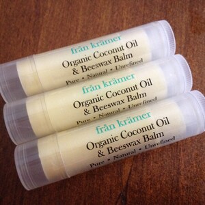 Pu&ograve; includere: Tre tubetti di balsamo per labbra con etichette bianche e tappi trasparenti sono disposti su una superficie di legno marrone. Le etichette riportano "fr&aring;n kr&auml;mer Organic Coconut Oil & Beeswax Balm". Il balsamo &egrave; beige chiaro.