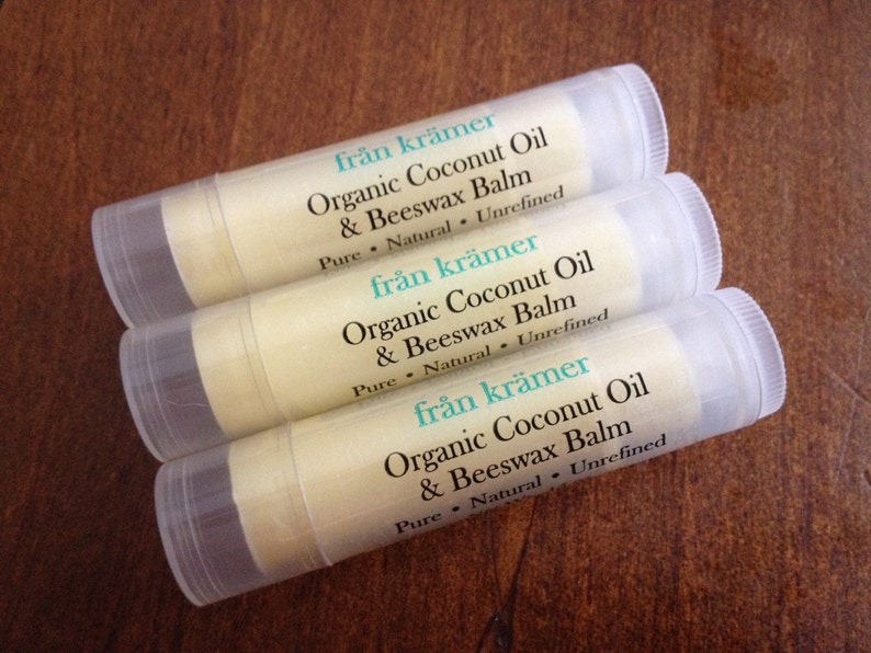 Pu&ograve; includere: Tre tubetti di balsamo per labbra con etichette bianche e tappi trasparenti sono disposti su una superficie di legno marrone. Le etichette riportano "fr&aring;n kr&auml;mer Organic Coconut Oil & Beeswax Balm". Il balsamo &egrave; beige chiaro.