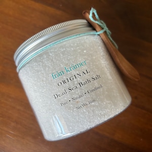 May include: A clear plastic jar filled with white Dead Sea bath salt. The jar has a silver lid and a light blue string tied around it, holding a small wooden spoon. Text on the jar reads "från krämer ORIGINAL Dead Sea Bath Salt Pure Natural Unrefined Net Wt. 16oz."