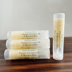 Pu&ograve; includere: Primo piano di quattro tubetti di balsamo per le labbra. I tubetti trasparenti hanno etichette bianche con la scritta "Organic Coconut Oil & Beeswax Balm". Il balsamo &egrave; di colore giallo chiaro. &Egrave; visibile anche il marchio "fr&aring;n kr&auml;mer".