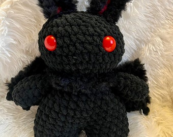 May include: A handmade black crochet plush toy in the shape of a moth. The plush has red button eyes, fluffy black fur accents, and small wings. The toy is sitting on a white, fluffy surface.