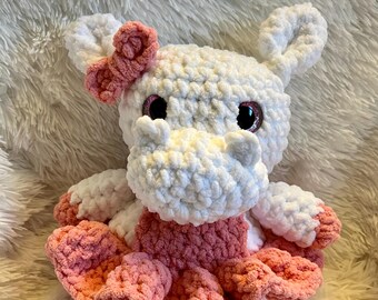 May include: A handmade crochet hippo plush toy. The hippo is white and pink, with a pink bow on its head and a pink dress. The toy has large, glittery eyes and is made with soft, textured yarn. A cute and cuddly stuffed animal.