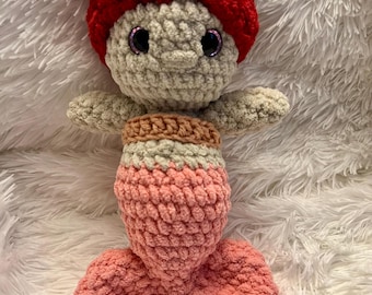 May include: A handmade crochet mermaid doll with a red cap and a flower accent. The doll has a light green face and arms, a brown and white top, and a pink tail. The doll is approximately 10 inches tall.
