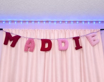 May include: A decorative banner featuring individual crocheted letters spelling the name MADDIE. The letters alternate between dark red and light pink yarn colors, suspended on a thin string against a solid light pink curtain background.