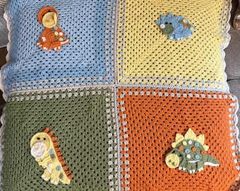 May include: A handmade crochet blanket featuring a patchwork design with four squares in blue, yellow, green, and orange. Each square has a dinosaur applique. The blanket has a white scalloped edge.