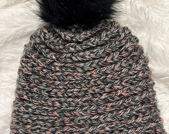 May include: A handmade knit beanie in gray, black, and peach tones, topped with a large black faux fur pom. The hat features a textured stitch pattern and a small leather tag with text.