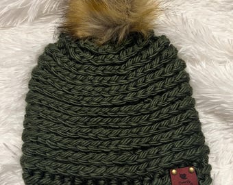 May include: A dark green, hand-crocheted beanie with a fluffy, light brown faux fur pom-pom on top. The hat has a ribbed texture and a small, burgundy leather tag with gold rivets. The tag reads "Crochet by Cozy B".