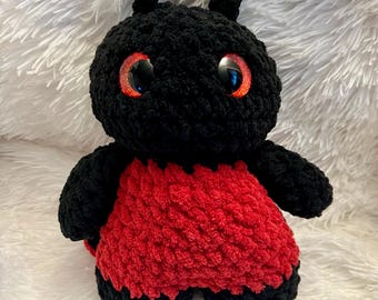 May include: A handmade black and red crochet bug plushie with large, reflective orange eyes. The plushie has a black head and limbs, with a red body. The plushie is sitting on a white, fluffy surface.