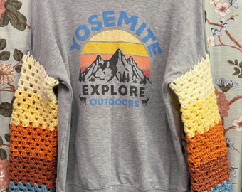 May include: Gray sweatshirt with crocheted sleeves. The sweatshirt features the word "YOSEMITE" in blue, a sunset graphic, and the words "EXPLORE OUTDOORS". The crocheted sleeves are striped with yellow, orange, blue, and gray.