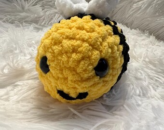 May include: A handmade, plush bee toy with a yellow body, black stripes, and white wings. The bee has large, black eyes and a smiling mouth. The toy is made of soft, textured yarn.