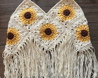 May include: Cream-colored crochet top with sunflower designs in yellow and brown. The top features a halter neck and a fringe along the bottom. The sunflowers are evenly spaced across the front of the top, creating a summery, bohemian look.