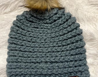 May include: A teal-colored, chunky knit beanie with a faux fur pom-pom. The hat features a ribbed design and a small, brown leather tag with a heart emblem. The pom-pom is a light brown color and adds a touch of flair to the winter hat.