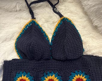 May include: A handmade crochet top in navy blue with a floral design. The top features a halter-style neckline and a bandeau-style bottom. The floral design incorporates teal, yellow, and red colors, creating a vibrant contrast against the navy blue.