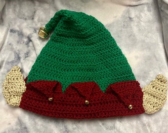 May include: A handmade crochet elf hat in green and red, with beige ear accents and a gold bell. The hat is designed with a pointed top and decorative gold bells on the brim, perfect for holiday celebrations.