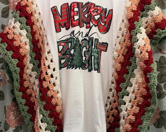 May include: White t-shirt with the words "MERRY and BRIGHT" in red, green, and white. The sleeves are a crocheted granny square design in red, green, peach, and cream colors. The shirt is hanging on a hanger.