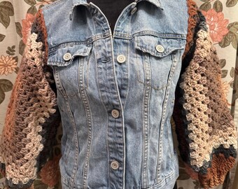 May include: A light blue denim jacket with crochet sleeves. The sleeves feature a gradient of brown, tan, and gray tones. The jacket has a classic collar, button-down front, and two chest pockets.