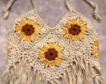 May include: Cream-colored crochet halter top featuring three sunflower designs with yellow and brown petals. The top has a fringed bottom and cream-colored ties. The crochet work is detailed and the sunflowers are centered.